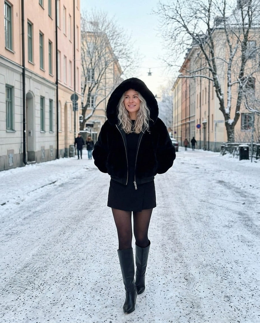Stina, fuskpälsjacka med huva för dam i svart, stylinginspiration på en snöig gata